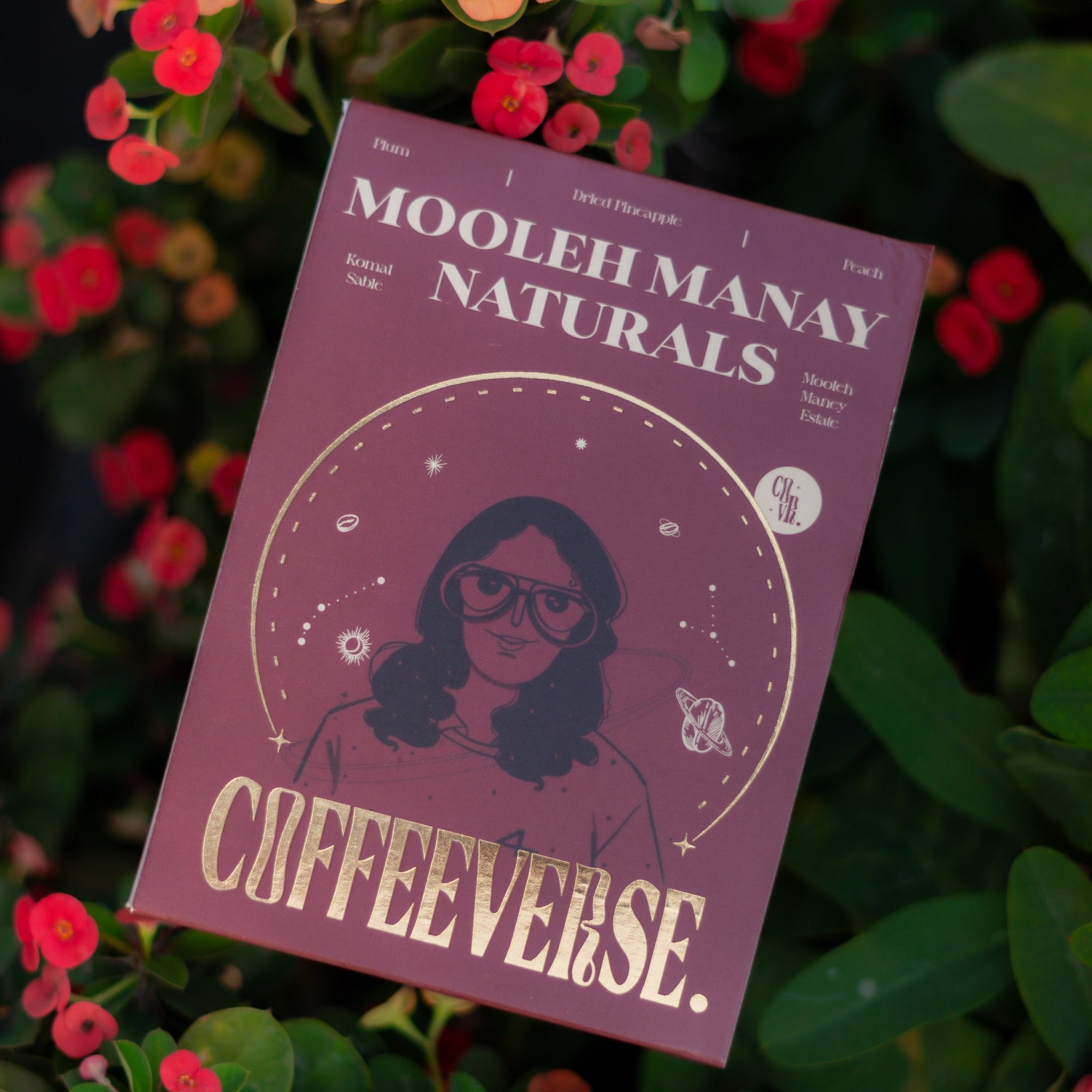 A pack of Mooleh Manay Naturals coffee placed on a surface with red and yellow flowers around it.