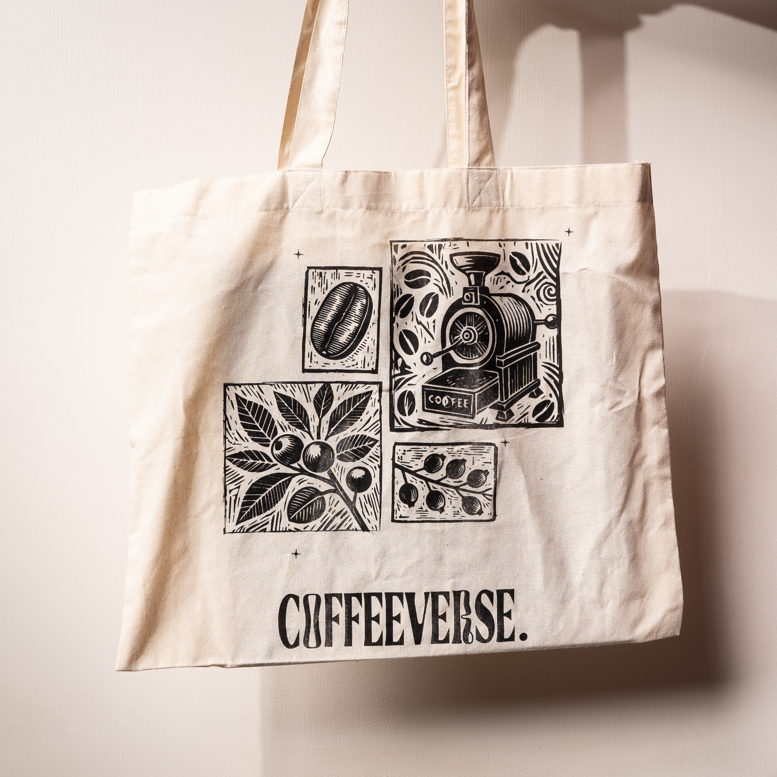 Beige Coffeeverse tote bag with black illustrations of coffee beans, plants, and a coffee roaster printed on the front.