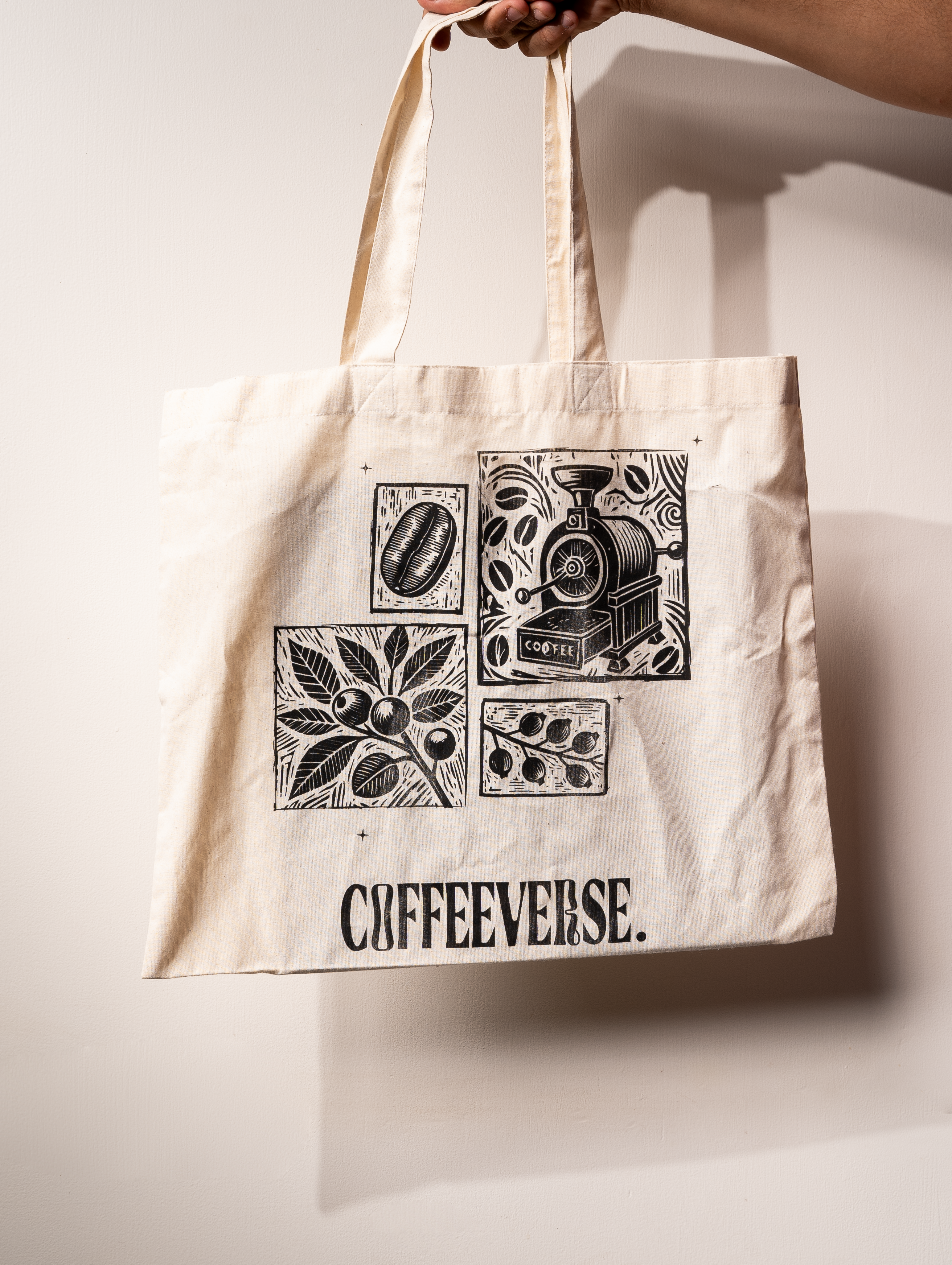 Beige Coffeeverse tote bag with black illustrations of coffee beans, plants, and a coffee roaster printed on the front.